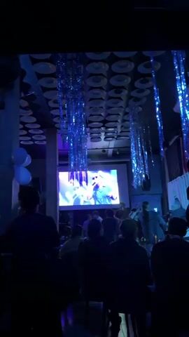 The video shows a dimly lit indoor venue, likely a club or bar, with a large projection screen at the front displaying a man singing. The screen is illuminated with bright blue and white lights, creating a stage-like atmosphere. Several rows of people are seated or standing in front of the screen, facing it. Some are silhouetted against the light, while others are more visible in the dim lighting. Blue and silver tinsel-like decorations hang from the ceiling. To the left of the screen, two white balloons are visible. The overall ambiance is that of a live performance or viewing party.