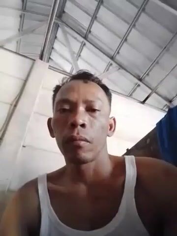 A man wearing a white tank top is looking at the camera from a low angle. He has short, dark hair and his face is the main focus. Behind him, a corrugated metal ceiling is visible, with white walls to his left and right. The lighting appears to be natural and somewhat dim.