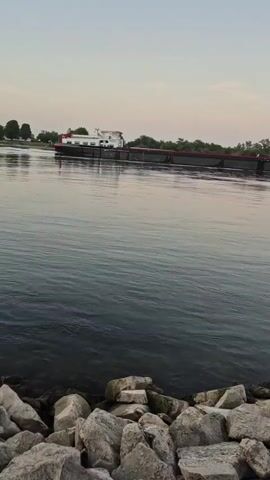 A barge with a white cabin structure and a long, dark hull with red trim travels down a wide river. The water is dark and calm, with small ripples. Rocks line the near bank of the river. Trees and a pale sky are visible in the background.