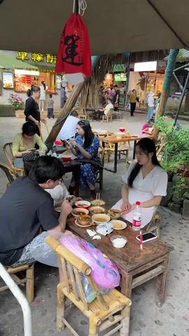 The video shows a group of people dining outdoors at wooden tables. Several dishes of food are placed on the table where a man and two women are seated. The man is eating while looking at his food. The woman to his right is looking down at her phone. The other woman sits next to her, looking straight ahead with her arms folded. Another table can be seen behind them, with a woman in blue looking at her phone and another woman looking at her phone to her left. An attendant is seen walking around, picking up dishes at tables. The background of the video appears to be a street food market, with several small shops and food stalls visible. The camera then moves towards the right, briefly showing another table with a man and woman seated before becoming blurry and focusing on nearby foliage.