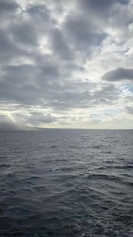 The video shows a view of the ocean from a boat. The sky is cloudy and the sun is breaking through the clouds in some areas, casting rays of light onto the water. The ocean is dark blue and appears to be choppy with small waves. In the distance, there is a coastline with hills or mountains. The camera pans slowly across the scene from left to right, revealing more of the coastline as it moves. There are no people or man-made objects visible in the frame other than what appears to be a distant ship on the horizon.