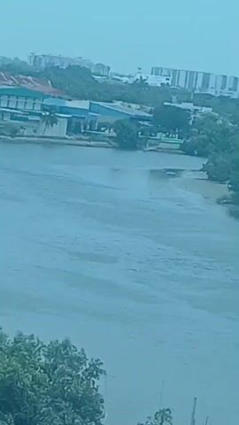 The video shows a view of a wide, murky river with several barges visible. On the riverbank, there are buildings, trees, and what appear to be industrial structures. The sky is overcast. The video progresses showing different angles of the industrial area and the river. There are no people visible in the video.