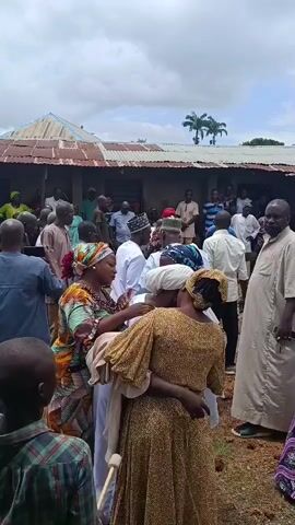 The video shows a gathering of people outdoors, with a building in the background. Many individuals are dressed in traditional clothing, with some wearing head coverings. In the foreground, two women are embracing, with one of them appearing to be comforted by the other. Other people are standing around them, some looking towards the embracing women, while others appear to be engaged in conversation or observing the scene. The environment appears to be somewhat rural, with dry ground and some vegetation visible. The sky is overcast. The overall mood seems somber, suggesting a moment of mourning or deep emotional expression.