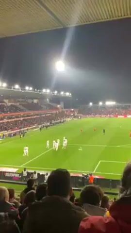 The video shows a soccer game at night, filmed from an elevated seating area. The stadium is packed with spectators, mostly dressed in red, and bright floodlights illuminate the green playing field. Players in white uniforms are positioned on one side of the field, facing players in red uniforms. People in the foreground, also likely spectators, are partially visible in the lower portion of the frame. Some are looking at the field, and one person appears to be using a cell phone. The background shows stadium seating filled with people, and the bright sky.