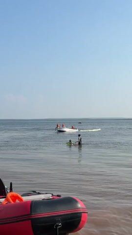 The video shows a body of water, possibly a lake or river, under a clear blue sky. In the foreground, the front of a red inflatable boat is visible on the sandy shore. In the middle ground, several people are enjoying recreational activities in the water. A small white boat with four individuals wearing orange life vests is in the background. Closer to the shore, two people are interacting in the water; one person is standing and appears to be assisting another person who is floating on a green inflatable ring. Further in the distance, two more small boats are visible on the water, creating small wakes. The water is calm with gentle ripples.