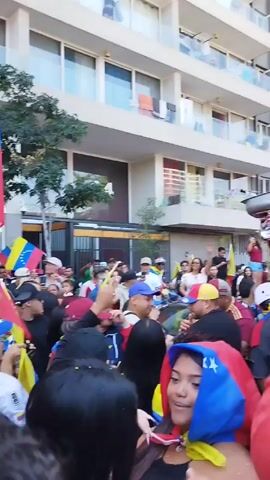 The video captures a lively street scene featuring a crowd of people waving Venezuelan flags. The atmosphere suggests a celebration or demonstration. A car modified with a large, pink speaker system in its trunk is prominently displayed, blasting music. The crowd seems to be enjoying the music and participating in the event with enthusiasm. The backdrop includes buildings and overhead power lines. The video is shot from within the crowd, giving a sense of being part of the event.