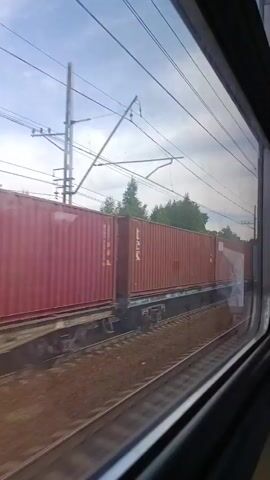 A freight train with multiple shipping containers is seen from a window as the train moves forward. The containers are different colors and brands, including red, gray, and orange ones. The train tracks are visible in the foreground, and trees and utility poles can be seen in the background. The sky is cloudy. The video appears to be taken during the day.