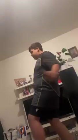 The video shows a young person in a gray shirt and black shorts in a living room. They are standing in front of a television set mounted on a wall and a small shelf containing books, photos, and a trophy above the TV. The person appears to be focused on something in their hands, possibly a remote or a small device. The camera angle is from below, looking upwards towards the person. The room is illuminated, but the lighting appears somewhat dim. The room includes other items such as what appears to be a green beanbag, a shopping bag, and a patterned rug, though these are mostly out of focus. The person is still and there is little to no apparent movement from the camera after the initial adjustment.
