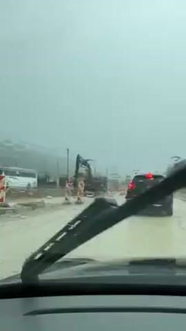 The view from inside a car shows a road in poor condition on a rainy day. The windshield wipers are operating. Ahead is a black car moving slowly through a muddy, wet construction zone marked by red and white striped traffic barriers. A yellow backhoe is visible on the left side of the road, along with a bus. Mountains are visible in the background.