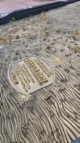 The video begins with a close-up view of a detailed relief map, possibly a historical or geographical representation, with intricate golden figures scattered across it. The map appears to be in an outdoor setting. The camera then pans up and away from the map to reveal a courtyard area with a large, golden statue of a person with arms raised in the air. Several people are visible in the background, walking around and observing the surroundings. The architecture suggests a religious or cultural site, with ornate buildings and structures in the background. The sky appears overcast with dramatic clouds. The video captures the scale and grandeur of the site, transitioning from the detailed map to the impressive statue and surrounding environment.