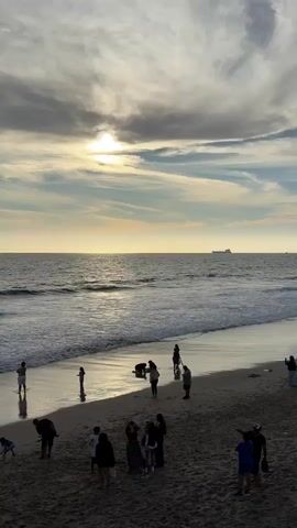 This video shows a beach scene during daytime. Several people are scattered across the sandy beach and near the water's edge. The ocean is visible with waves gently rolling in. In the distance, several cargo ships are sailing on the horizon. The sky is partly cloudy with the sun visible through the clouds, creating a soft, diffused light. The overall atmosphere appears relaxed and casual, with people enjoying the beach.