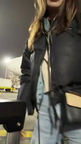 A woman in a black puffer jacket, light-wash jeans, and a backpack approaches a man in a white puffer jacket. They embrace and kiss on the cheek. The scene takes place outside at night, possibly at an airport, as indicated by the ground markings and the presence of an airplane boarding ramp in the background.