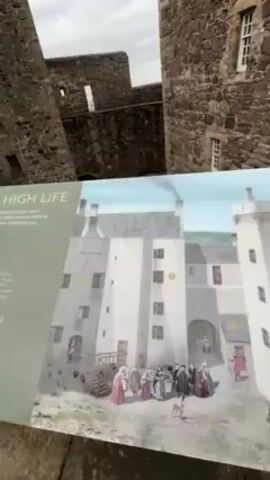 The video shows a person holding a sign that depicts a historical rendering of Blackness Castle in Scotland. The rendering shows people in historical clothing in the castle courtyard. The camera moves from the sign to the castle itself, showing the stone walls and towers. The sky is overcast.