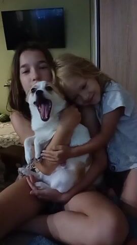 A teenage girl holds a small dog on her lap. A younger girl is leaning her head on the teenage girl's shoulder, hugging the dog. The two girls and the dog are sitting on the floor, with a television on the wall in the background.