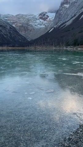 The video shows a clear, still lake, partially frozen, with a rocky shore. Snow-covered mountains rise in the background under a cloudy sky. Someone throws a rock onto the ice, causing a cracking sound.