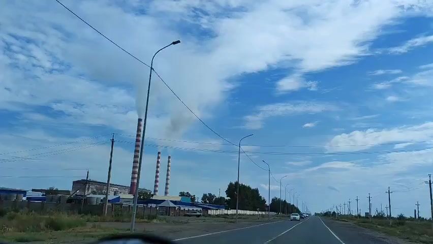 The video captures a view from inside a moving vehicle, looking out at a two-lane road. On the left side of the road, a large industrial facility is visible with several tall, striped smokestacks emitting white smoke. The facility appears to have a long, low-rise building attached to it. A fence surrounds the industrial area. Along the right side of the road, there are numerous utility poles with wires stretching across the scene. A few cars are driving in the same direction as the viewer, and the sky is blue with scattered white clouds. The overall environment appears to be somewhat industrial and rural.