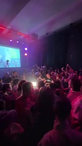The video shows a crowd of people at a nightclub. The room is dark with purple and red lighting. A DJ is at the front, behind a metal barrier. A projection screen behind the DJ shows a logo with “MUS3 YOU” and “ba day CLvb” around a heart. People are holding up phones, dancing, and interacting with each other. The setting appears to be a musical performance or club event.
