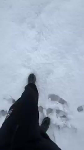 A first-person perspective video shows a person standing in snow. The person wears dark-colored pants and dark shoes. There are footprints in the snow.