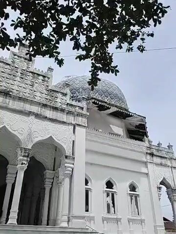 The video shows the exterior of a white building, likely a mosque or historical structure, with intricate architectural details. The building features arched doorways supported by pillars, and decorative patterns on the facade. A dome-shaped roof is visible on one section. Part of the building's roof appears to be undergoing repair or construction. A minaret can be seen in the background. The foreground includes foliage, suggesting a natural setting. The sky is overcast.