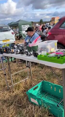 The video pans across a flea market or car boot sale set up in a grassy, outdoor area under a cloudy sky. In the foreground, a white folding table is laden with fishing equipment. Several fishing reels, some in black and silver, are displayed prominently. Behind the table, a person wearing a blue t-shirt and a black cap is visible, partially obscured by the table and equipment. Various fishing accessories, including tackle boxes and plastic containers filled with lures and other items, are arranged on the table. Further back, a white car is parked, and more stalls and tents are set up. In the background, other people can be seen browsing and interacting at the stalls. The overall impression is of a busy outdoor market selling fishing gear.