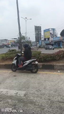 A person wearing a black hoodie and a helmet is sitting on a silver scooter, stopped next to a street. The person is wearing red shoes. The scooter has a license plate. The background shows a street with traffic, including cars and other scooters. There are buildings and billboards visible. The sky is overcast. The video appears to be shot from a moving vehicle, possibly a car, given the slight camera shake and the perspective. The time stamp at the bottom indicates "TECNO POVA 7".