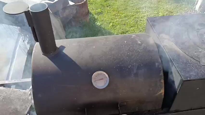A close-up aerial shot shows racks of ribs smoking on a grill. The ribs are seasoned and appear to be in various stages of cooking. The grill is outdoors, with grass and a shed visible in the background. Smoke is rising from the ribs and the grill. A thermometer is visible on the side of the smoker.