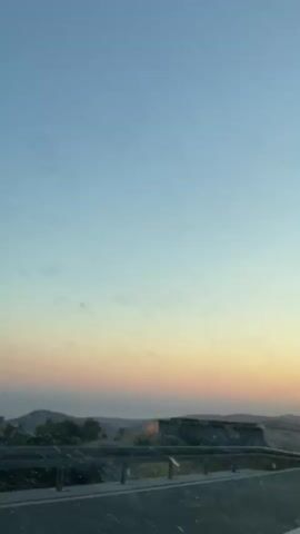 The video shows a view from a moving vehicle traveling along a road. The sky is clear and transitioning from a pale blue to soft orange and pink hues, suggesting it is either sunrise or sunset. There is a guardrail along the side of the road. In the distance, there are hills and possibly the sea. On the left side of the road, there is a stone structure. In the middle of the video, a silver car is visible parked by the guardrail, and two people are standing near it. Further down the road, another silver car is seen passing by in the opposite direction. A road sign is also visible. The perspective suggests the video is taken from the driver's or front passenger's seat.