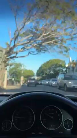 The video, filmed from the interior of a car, presents a view of a street under a clear, blue sky. The street is lined with trees casting shadows, and parked cars occupy both sides. In the distance, one can discern a body of water, possibly the sea or a bay, hinting at a coastal or near-coastal location. The camera's perspective is from the driver's seat, capturing the dashboard and steering wheel, with gauges and controls visible. The car appears to be moving slowly or at a standstill due to traffic conditions. The video captures a calm, sunny day scene, likely during daytime hours.