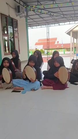 A group of seven young girls, all wearing headscarves, sit cross-legged on a light-colored tiled floor under an outdoor awning. They are playing round, hand-held frame drums. The girl in the front row on the left has a light brown drum, the girl next to her has a similar drum. The girl in the second row, left of center, also has a light brown drum. The girl to her right has a darker drum. The girl in the front row, right of center, has a light brown drum. The girl behind her is also holding a drum. The girl furthest right is partially visible and appears to be holding a drum. They are all looking towards the front of the frame. In the background, there is a building with green trim, a white wall, and windows. There are also colorful triangular flags strung across the awning. Beyond the building, there is a clear sky and some vegetation. The lighting suggests it is daytime.