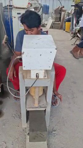 A man in a dark blue shirt and red pants is crouched next to a concrete testing machine in an outdoor area with concrete debris on the ground. The machine has a white housing on top, a metal frame, and a visible screw mechanism. A grey concrete block is placed on the lower part of the machine, and the machine's mechanism is positioned above it. In the background, a blue barrel, a yellow warning sign, and other construction materials are visible. Another person in the background is wearing brown boots and dark pants. The man is looking at the machine and appears to be operating it. The machine is in operation, exerting pressure on the concrete block.