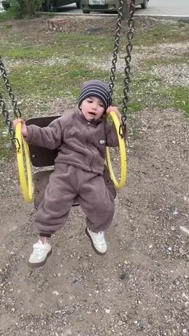 A young child, likely a toddler, is seated on a playground swing. The child is wearing a warm, brown fleece tracksuit, a striped knit hat in shades of blue, brown, and white, and light-colored sneakers. The swing features a brown wooden seat and yellow plastic-covered handles or chains, which are attached to metal chains.

The video shows the child gently swinging back and forth, vocalizing with "baba" sounds. A distinct green substance is visible on the child's chin and around the mouth area.

The scene takes place outdoors during the daytime, under what appears to be an overcast or cool sky, as suggested by the child's warm clothing. The ground beneath the swing is a mix of dirt and gravel, with scattered patches of green grass. The background, largely out of focus, reveals more grassy areas and a distant tree trunk. A small, indistinct emblem or patch is visible on the left chest of the child's tracksuit. The provided location context for this media file is Sol-Iletsk, Russia.