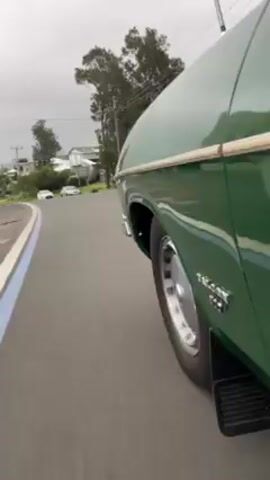 The video shows a green car driving on a paved road with a blue line on the side. The car's tire, wheel, and mudflap are visible. The car appears to be turning to the left. In the background, there are houses, trees, and a grey sky. The word "FALCON 500" is visible on the car's side. The camera angle is low and from the side of the car.