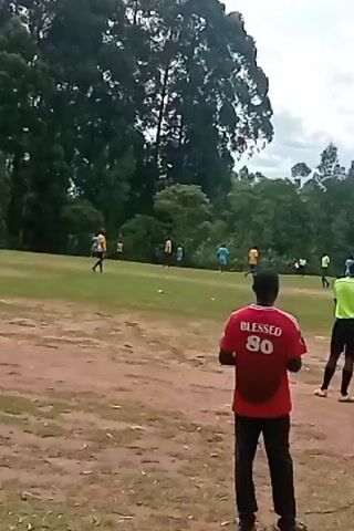 The scene appears to be a soccer or football match in progress on a field with patchy grass and exposed dirt patches. Several players are visible, wearing different colored jerseys - primarily blue and orange. One individual is wearing a bright yellow shirt, possibly a referee, holding a flag. In the foreground, a person wearing a red shirt with the words "BLESSED 80" is watching the game. Another person is sitting on the ground to the right of the person in the red shirt, also observing the match. The background features lush green trees and a sky with scattered clouds.