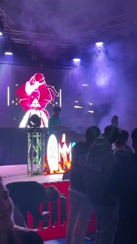 The video shows a DJ performing at a concert or club. The DJ is standing behind a table with equipment, in front of a large screen displaying a graphic. The lighting is colorful with fog or smoke. A crowd of people is watching the performance. A hand is raised at one point.