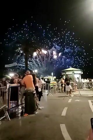 The video shows a nighttime outdoor scene with fireworks illuminating the sky. A crowd of people is gathered, some standing behind metal barriers, watching the display. In the background, there are lights from buildings and possibly vehicles. The fireworks are burst into various shapes and colors, creating a festive atmosphere. One person in the foreground is holding up a phone, possibly to record the event.