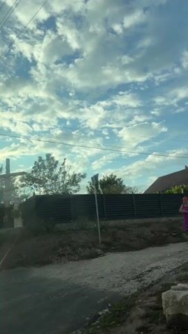 The video begins with a view of a cross with a figure of Jesus on it, situated in front of a fence and trees. The camera then pans to the right, showing a field of corn and a utility pole. The scene continues to pan, revealing more trees, another utility pole, and a wooden structure in the distance. The sky is filled with clouds and sunlight. The overall environment appears to be rural.