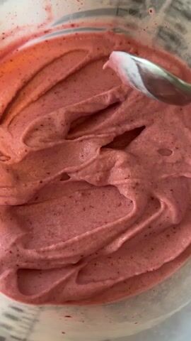 The media file is a close-up, overhead shot of a bowl filled with a pink, creamy substance, likely a dessert such as sorbet or ice cream. The texture is smooth with small flecks of red visible throughout, suggesting it might be berry-flavored. A spoon is actively swirling through the mixture, creating wavy patterns and highlighting the creamy consistency. In the background, faint markings on a transparent container suggest measurement lines, possibly indicating a blender jar or a serving bowl. The lighting is bright, and there are no discernible shadows, suggesting an indoor setting with artificial light or a well-lit environment. The scene is static, focusing solely on the preparation or serving of the dessert. There is no clear indication of the time of day, weather, or specific location beyond the container's markings. The overall mood is neutral, with the focus being on the texture and visual appeal of the food. No visible text is present in the media.