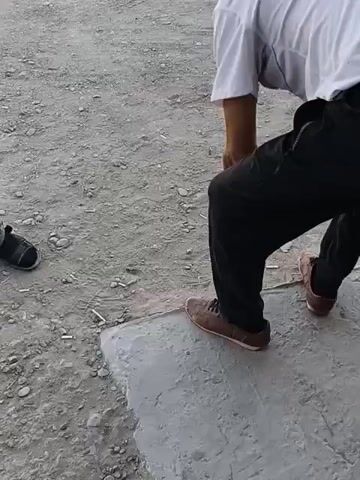 The video begins by showing a person's legs and feet from a low angle, with the person wearing dark pants and brown shoes. The ground is a mixture of dirt and gravel. In the first frame, the person is holding a small, dark object in their right hand. In the subsequent frames, the person's hands are not visible. Another person's legs and feet, wearing black shoes, are visible on the left side of the frame in the first two frames. In the third and fourth frames, another person is visible from the waist down, wearing dark pants and holding a can and a cigarette in their right hand. A plastic water bottle is on the ground. The background remains the dirt and gravel environment.