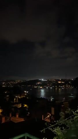 A nighttime aerial view of a city illuminated by numerous lights. A body of water, likely a river or strait, is visible in the middle ground, reflecting the lights of the city. Boats can be seen on the water. In the foreground, the silhouettes of houses with red tiled roofs are visible, along with some foliage. The sky is dark with scattered clouds.