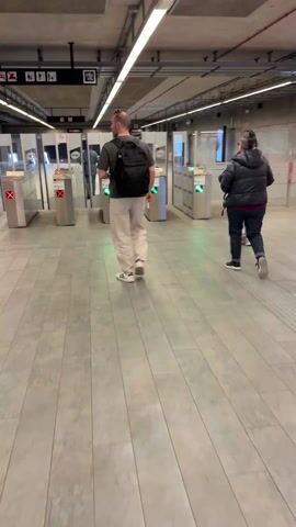 The video shows three individuals walking through a subway station. They are using automated ticket gates to pass through a barrier. The first person, a man, is wearing a t-shirt, light-colored pants, and white sneakers with black stripes. He carries a black backpack. The second person, also walking through the gate, is wearing a dark jacket over a red and black plaid shirt, along with dark jeans and sneakers. The third person wears light wash jeans, and a dark vest over a white top. The gates are metal with green lights indicating passage. The subway station has a gray tile floor and a series of fluorescent lights overhead. The overall impression is that of a modern, functional, and well-lit transit environment.