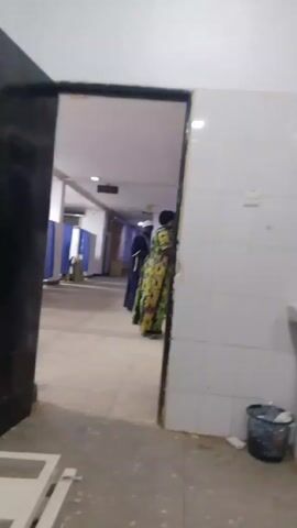 The video shows a hallway in a hospital. The hallway has white tile walls and a ceiling with recessed lighting. On the left of the hallway, there are blue privacy curtains. Further down the hall, two individuals stand: a man dressed in a dark robe and white cap, and a woman in a bright, patterned dress. The woman is standing against the wall. The view is framed by a doorway, with the frame visible on the right.