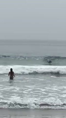 A man is surfing on a wave in the ocean while a woman stands in the shallow water near the shore. It appears to be a cloudy day.