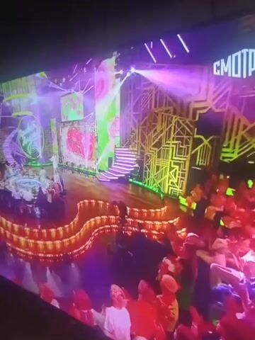The video begins with a wide shot of a brightly lit stage adorned with pink and green floral motifs and geometric gold structures. In the foreground, an audience is visible, their faces illuminated by the stage lights. As the video progresses, the camera zooms in on the stage. A woman with blonde hair, wearing a white top, stands center stage, holding a microphone and singing. She appears to be in the midst of a performance, with her eyes closed and her head tilted back. The background behind her is a vibrant red, also featuring abstract floral patterns. The overall ambiance suggests a live musical performance or a televised show.