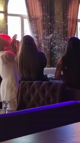 In Yablonovskiy, Russia, a group of people are gathered indoors, likely at a venue or restaurant. The scene is filled with bubbles, suggesting a celebratory or festive atmosphere, possibly a birthday party, wedding, or other special occasion. Several women are visible, with their backs to the camera for the most part. One woman in the foreground is seated in a plush, tufted armchair. Another woman, wearing a white outfit, is standing and appears to be interacting with the bubbles. The lighting in the room is dynamic, with colorful spotlights, including green and purple hues, casting a festive glow. Heavy, ornate curtains adorn the windows, and the overall decor suggests a somewhat formal or elegant setting. The time of day is not explicitly stated, but the indoor lighting and the presence of artificial lights suggest it could be evening or a dimly lit indoor space. No text is visible in the media.