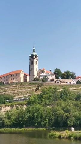 The video shows a scenic view of a church tower and buildings situated on a hillside overlooking a body of water. The hillside is covered with terraced vineyards and lush green trees. The sky is clear and blue. The water in the foreground is calm and reflects the scenery. The buildings appear to be made of stone and have red tile roofs. The church tower is tall and slender with a green, pointed spire. The scene suggests a tranquil and picturesque location, possibly in a rural or historical setting.