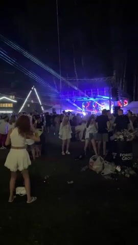 A night scene at an outdoor event with a stage and lights. A crowd of people are gathered, many appearing to be dancing. The camera focuses on a woman with red hair in a white outfit in the foreground, facing away from the camera and dancing. There is a trash can overflowing with garbage to her right. Laser lights are projected from the stage into the night sky.