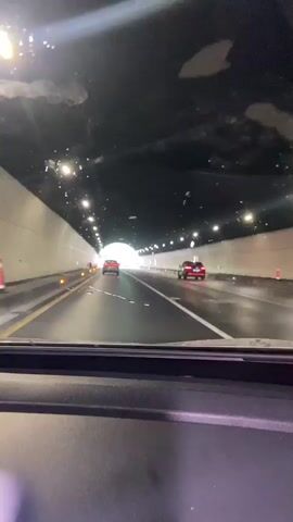 The video shows a view from inside a car driving through a tunnel. The tunnel has white walls and a dark ceiling with lights. There are two red cars visible ahead, driving in the same direction. The road appears to be wet, suggesting it might be raining or has recently rained. The view is from the front of the car, looking through the windshield.