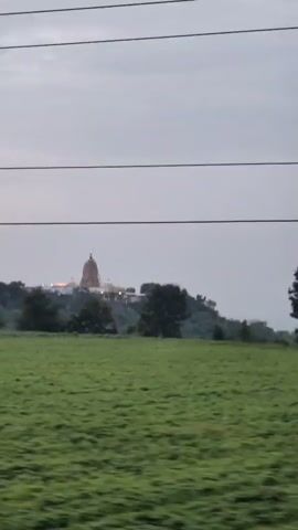 The video shows a landscape scene. In the foreground, there is a field of green crops. In the middle ground, there are trees and a hill. On top of the hill, there is a building with a dome, possibly a temple or shrine. Overhead, there are power lines. The sky is overcast. The video appears to be shot from a moving vehicle, as evidenced by the slight blur and the way the scene shifts from left to right.