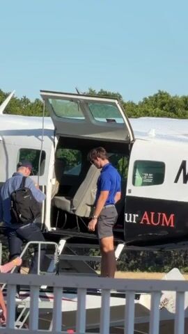 A group of people are boarding a small airplane. The plane is white with black and red lettering that says "urAUM". The rear hatch of the plane is open, and a set of stairs leads up to the opening. A man wearing a blue shirt and gray shorts stands by the opening, seemingly assisting with boarding. Another man in a blue shirt and a baseball cap is climbing the stairs, carrying a black backpack. To the left, another person in a light-colored shirt and khaki shorts is visible on the stairs. The sky is clear and blue, and trees are visible in the background. The scene appears to be taking place outdoors on a sunny day.