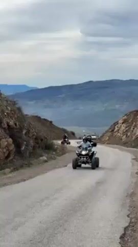 A video shows a group of ATVs driving on a winding mountain road. The road is paved, but shows signs of wear. The ATV in the foreground has two people on it, both wearing helmets. The surrounding landscape is mountainous, with a valley visible in the distance. The sky is overcast. The ATVs are moving away from the camera.